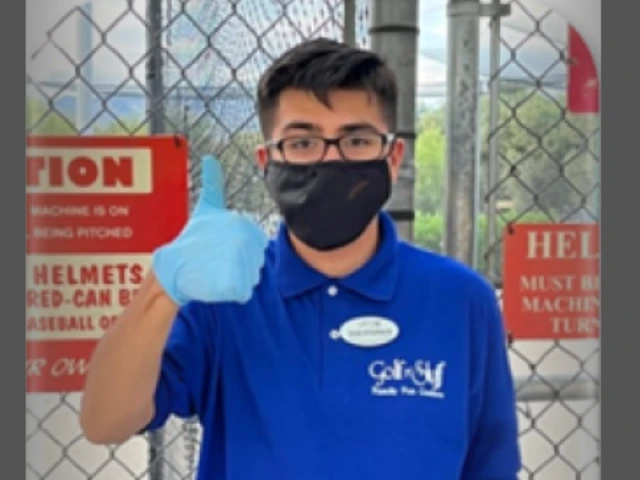 Young adult with short dark hair and glasses, wearing a blue shirt and a black medical mask, giving a thumbs-up to the camera.