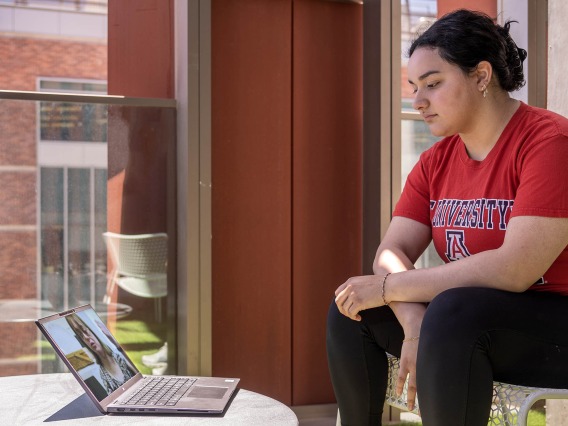 Student in a red University of Arizona shirt sitting with their hands crossed in front of them watching the interdisciplinary training session on their laptop.