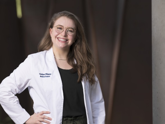 White woman with long light brown hair, wearing glasses and a white medical coat, smiling with her hand on her hip