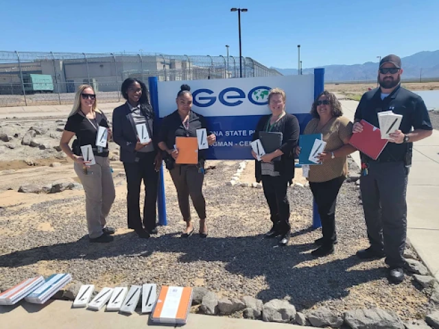 Six people holding LiveScribe pens and standing outdoors in front of a GeoGroup sign, with chain link fence and prison facility visible in the background