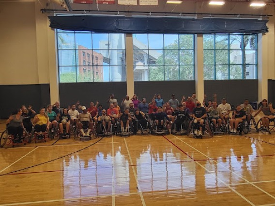 A large group people in wheelchairs posed in front of gym windows 