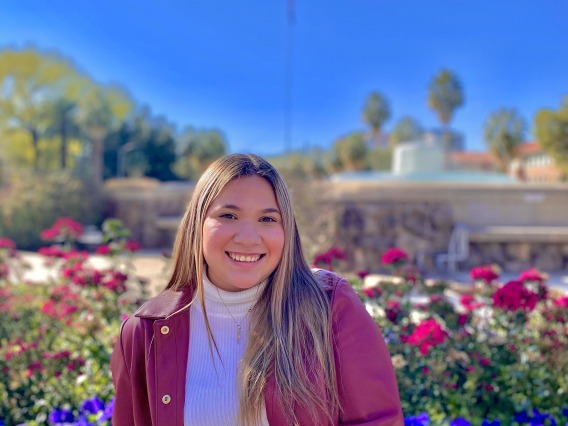Paulette is wearing a white turtle neck shirt with a red jacket and jeans. Paulette's hair is blonde with brown roots, she is light-skinned and is smiling. Paulette is sitting down in front of a water fountain with flowers in the background during a sunny day.