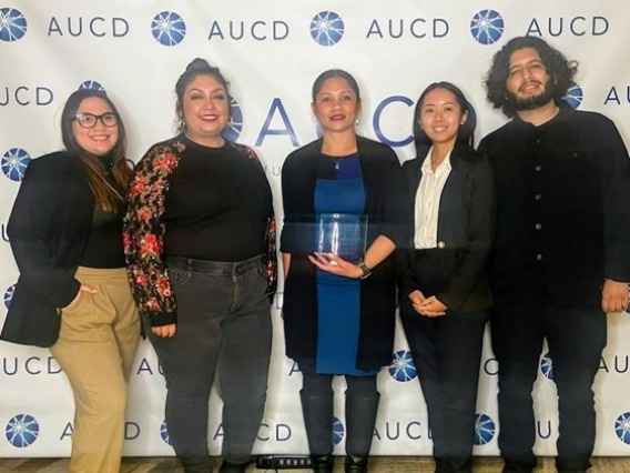Sonoran Center AUCD Conference attendees standing in front of a wall with the AUCD logo on it. Left to right: Paulette Nevarez, Celina Urquidez, Jacy Farkas, Nadine Chau, Abraham Venegas