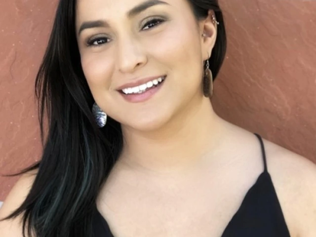 Zonnie Olivas, young Native woman in a black dress standing in front of a brick wall and smiling