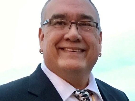 Jim Warne, a Native American man wearing a black jacket, white shirt, and brown tie