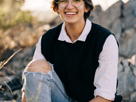 student sitting on rocks in front of desert background, wearing a white collared shirt with lack vest and jeans, swearing glasses with short brunette hair smiling at the camera