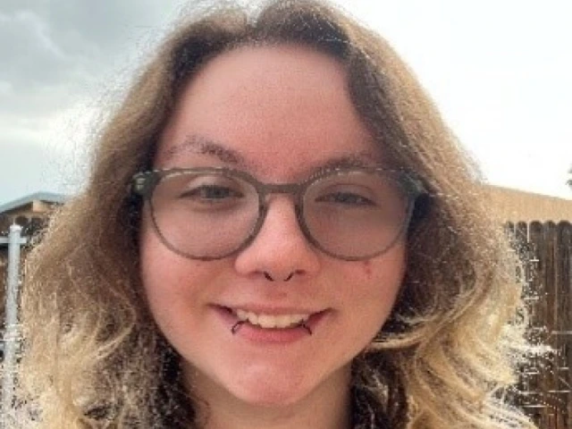 Elexander Childers, a young person with shoulder-length blonde hair and glasses, wearing a collared shirt, standing outside and smiling