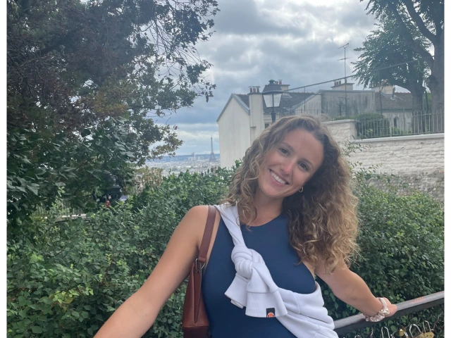 Photo of woman in front of a grassy landscape, wearing a navy blue top and white skirt, with blonde curly hair, smiling at the camera