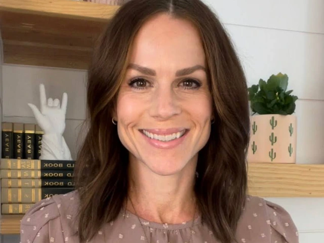Amber Hansen, a woman with long brown hair, wearing a lavender top with white spots and a gold necklace, standing in front of a shelf and smiling. 