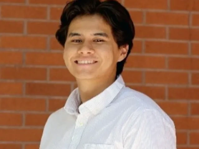 Joseph Fraire, a young person with dark hair wearing a white button-down shirt, standing in front of a brick wall and smiling