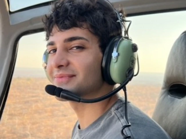 Photo of young man wearing a blue grey long sleeve shirt, has pilot headphone on and is in a helicopter. He has brown curly short hair and is smiling at the camera