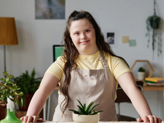A woman with autism with long brown hear standing in front of a potted plant smiling.