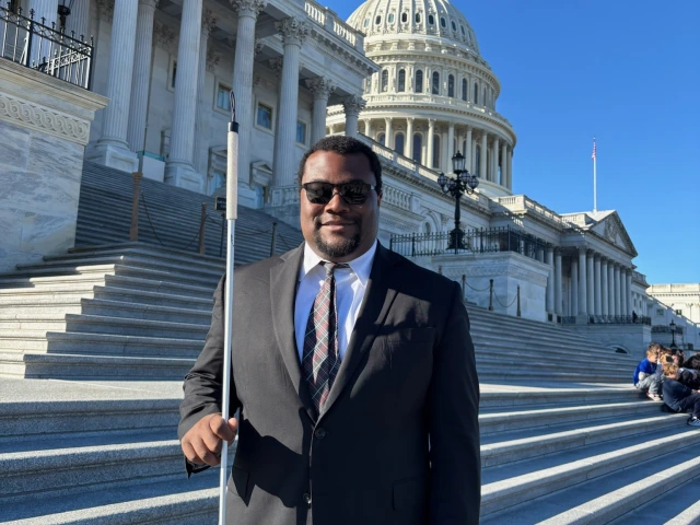 Jordan Moon, a man with dark skin and short hair, wearing a suit and sunglasses and holding a cane, standing in front of the US Capitol Building