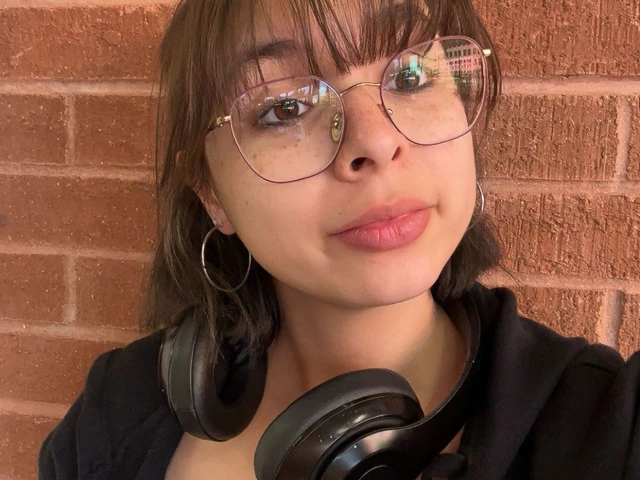 photo of Ariana with brown hair and bangs, wearing a black sweater and glasses with rose gold rims, is staring at the camera