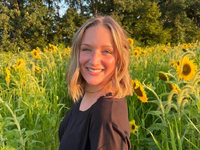 woman with short brunette hair, she is wearing a black blouse and standing in front of a sunflower field smiling at the camera