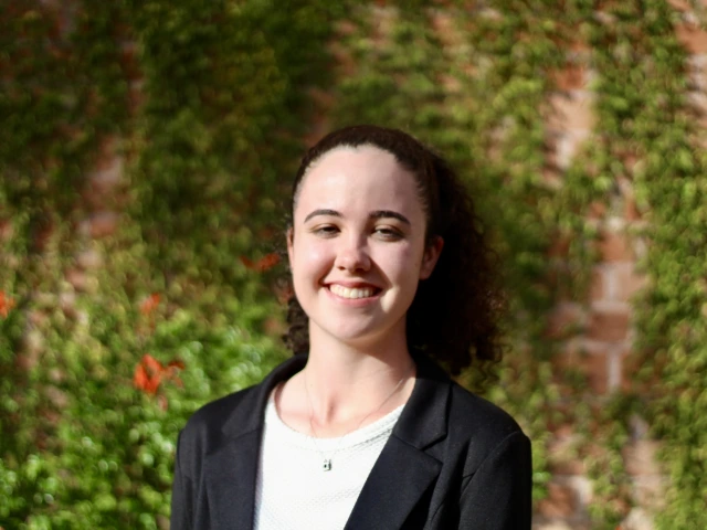 photo of young woman with curly hair in a pony tail, she is wearing a white shirt and black blazer and is smiling at the camera