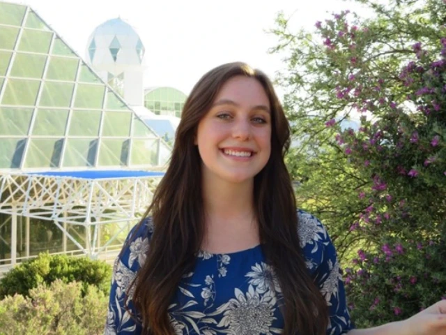 Nicole DiGonno, a Caucasian girl with brown hair and blue eyes stands smiling in front of Biosphere 2 in Tucson, Arizona. She is wearing a blue blouse with white flowers