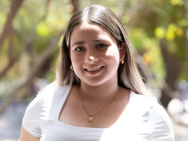 Ana Fernandez, a woman with light skin and blonde hair, wearing a white top, standing outside near some trees