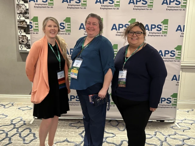 Jessica Winchester (left) and Rosa Rojas Franco (right) pose in front of a banner with the logo of the APSE Employment First, Employment for All Conference. 