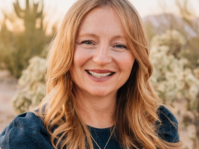 Woman wearing a navy blue shirt, has long strawberry blonde hair and she is smiling at the camera