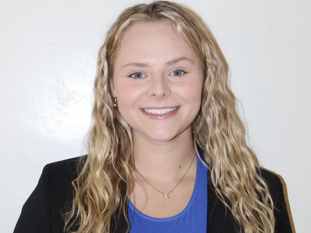 photo of young woman wearing a blue shirt and black blazer, she has blonde wavy hair and is smiling at the camera