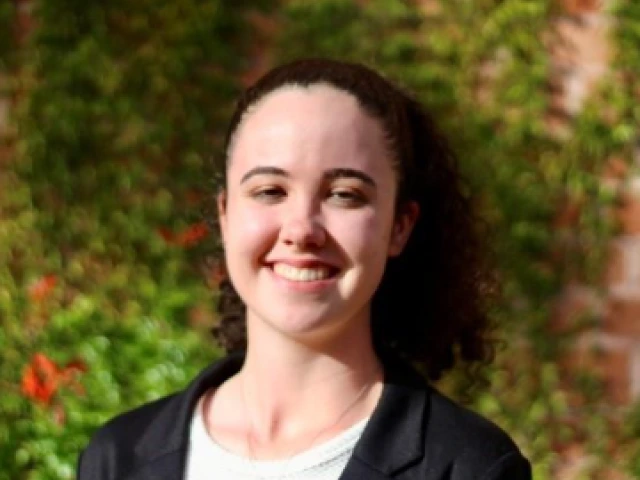  Smiling young woman with curly brown hair pulled back in a ponytail, wearing a black blazer over a white top, standing outdoors in front of a leafy green background 