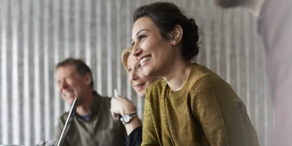 An adult woman in a yellow sweater, with brown hair pulled into a loose bun, sitting in a lecture and smiling.