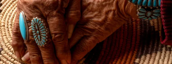 Elder hands wearing turquoise jewelry, clasped on top of a woven basket