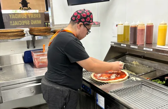 A person in a kitchen, adding toppings to a pizza.