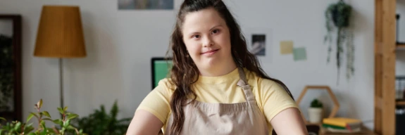 A woman with autism wearing a yellow shirt and has long brown hair. She is planting a potted plant and smiling.