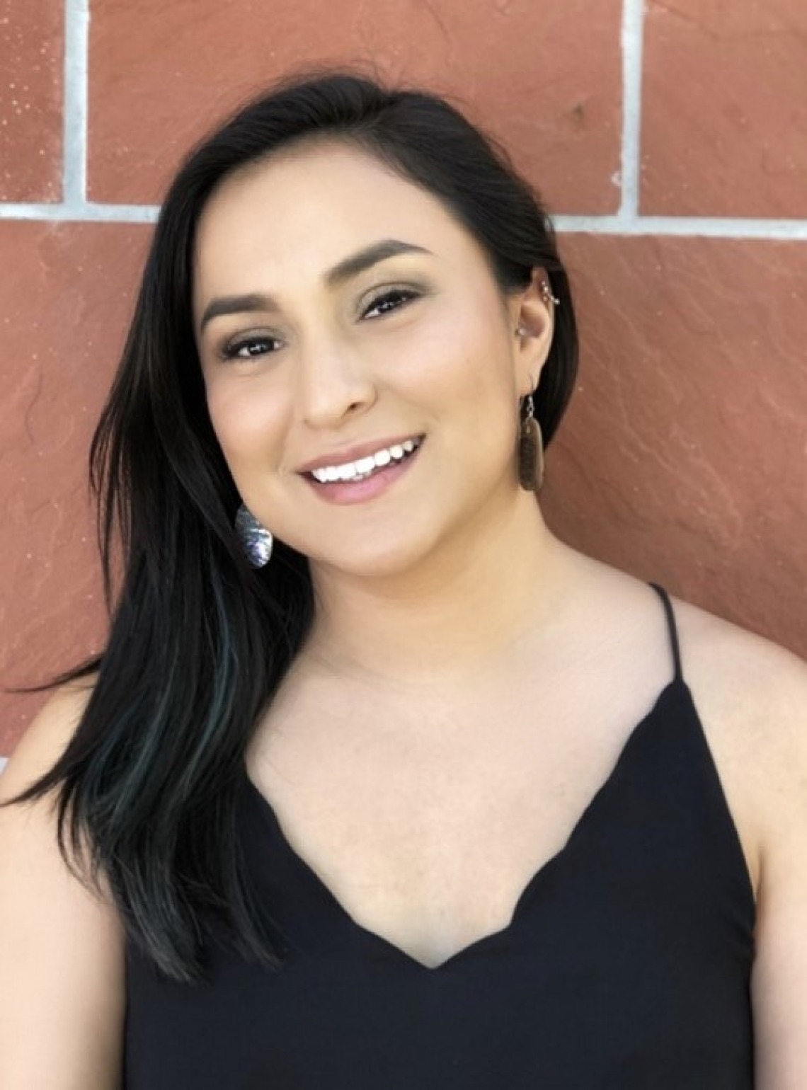 Zonnie Olivas, young Native woman in a black dress standing in front of a brick wall and smiling