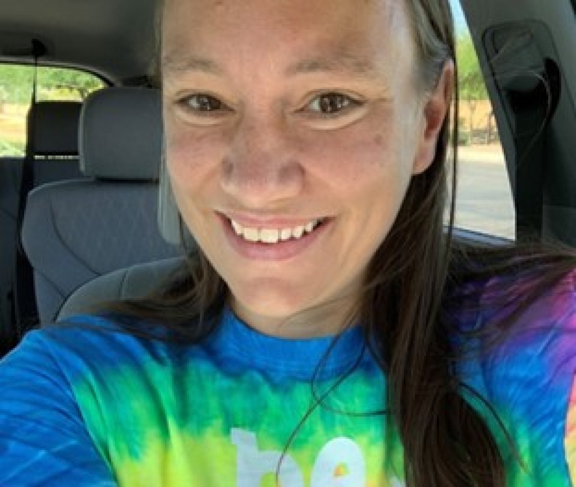 Katie Kwiatkowski, a woman with long brown hair, wearing a tie-dye shirt, smiling and sitting in a car