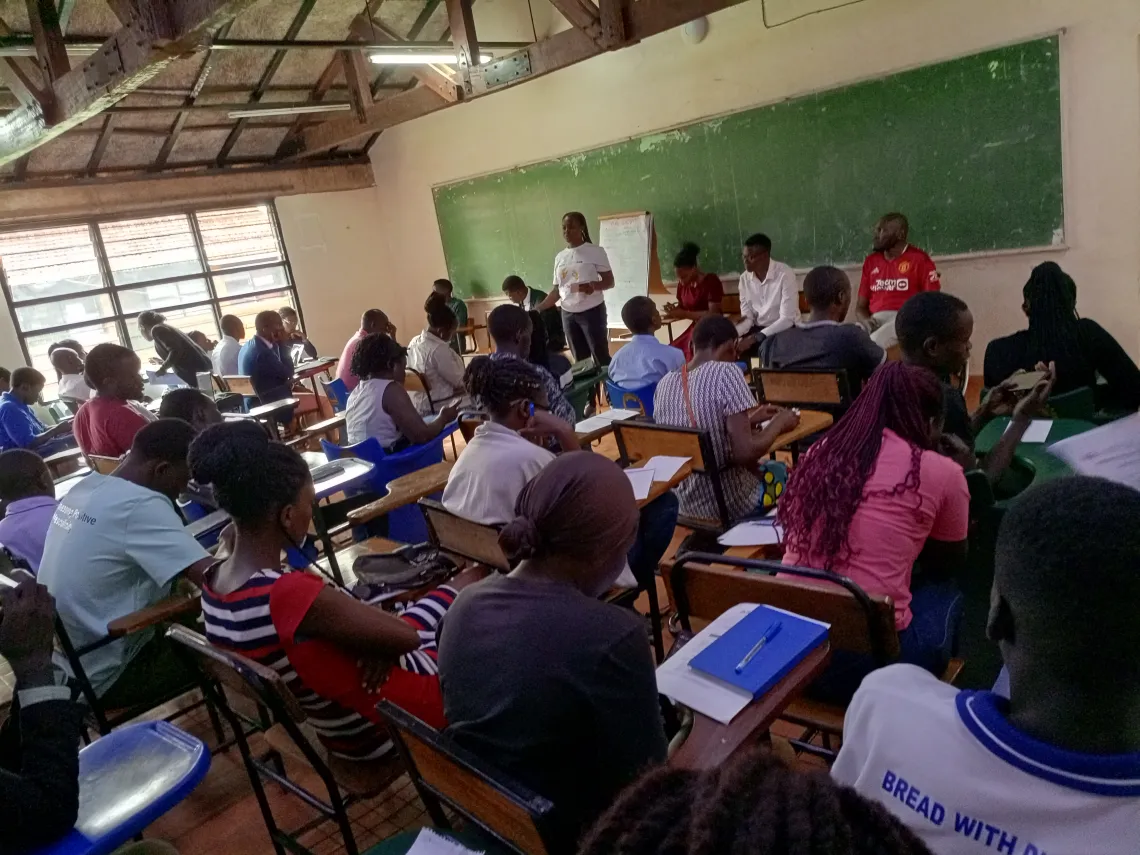 Crowded classroom full of students facing towards a wall with a chalkboard and a man with a red shirt sitting in a wheelchair.