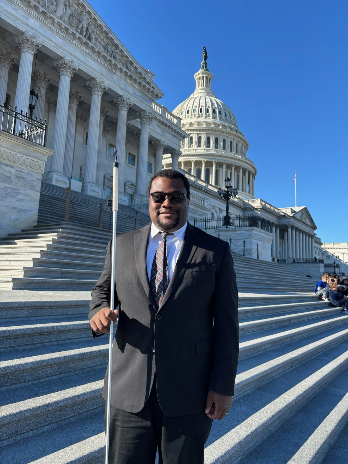 Jordan Moon, a man with dark skin and short hair, wearing a suit and sunglasses and holding a cane, standing in front of the US Capitol Building