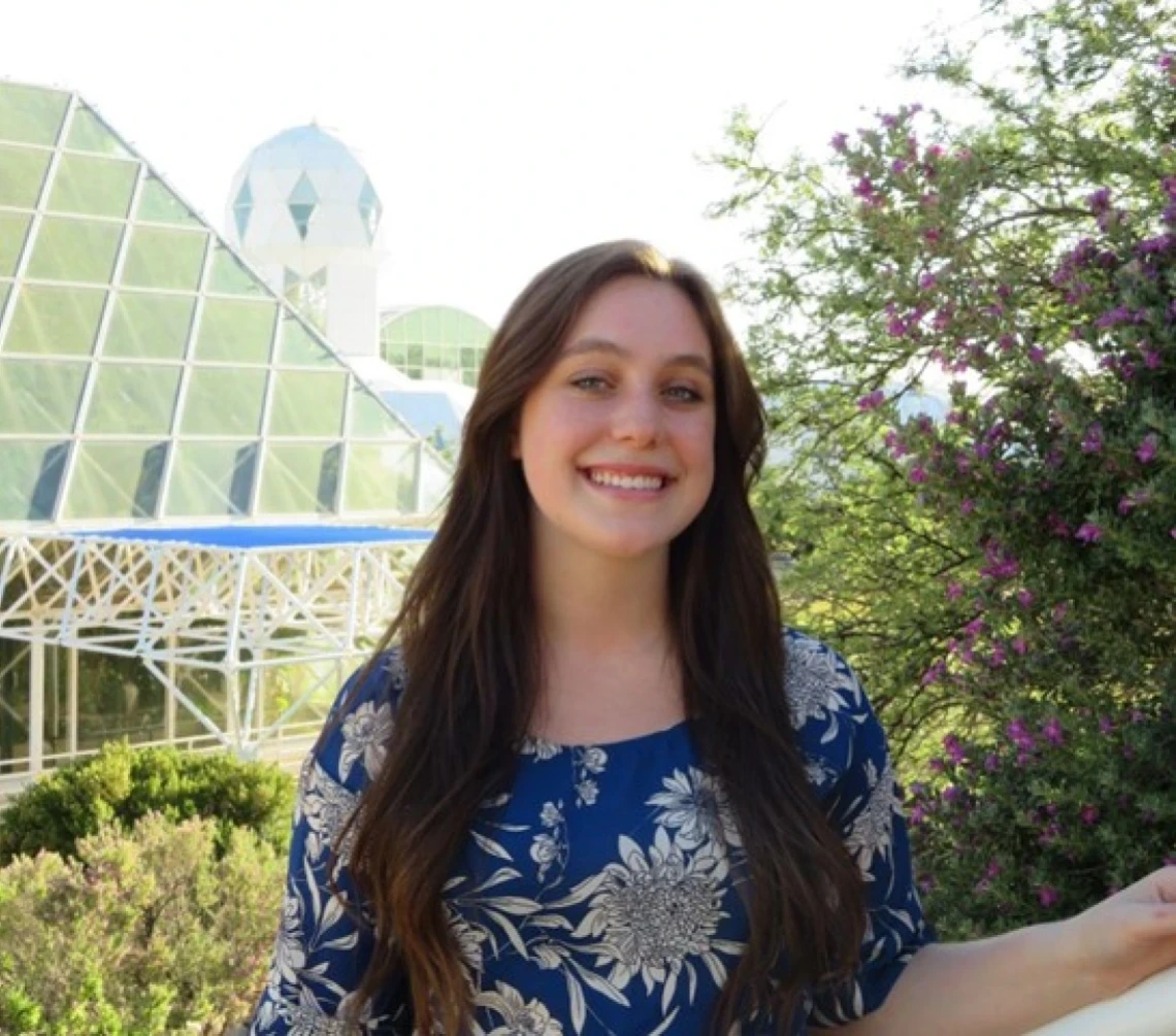 Nicole DiGonno, a Caucasian girl with brown hair and blue eyes stands smiling in front of Biosphere 2 in Tucson, Arizona. She is wearing a blue blouse with white flowers