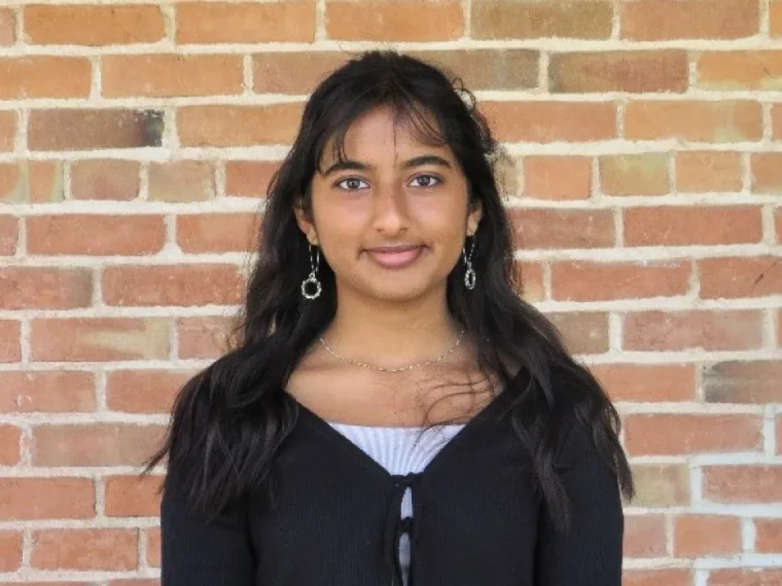 Esha Mahur, wearing a black top, standing in front of a brick wall and smiling
