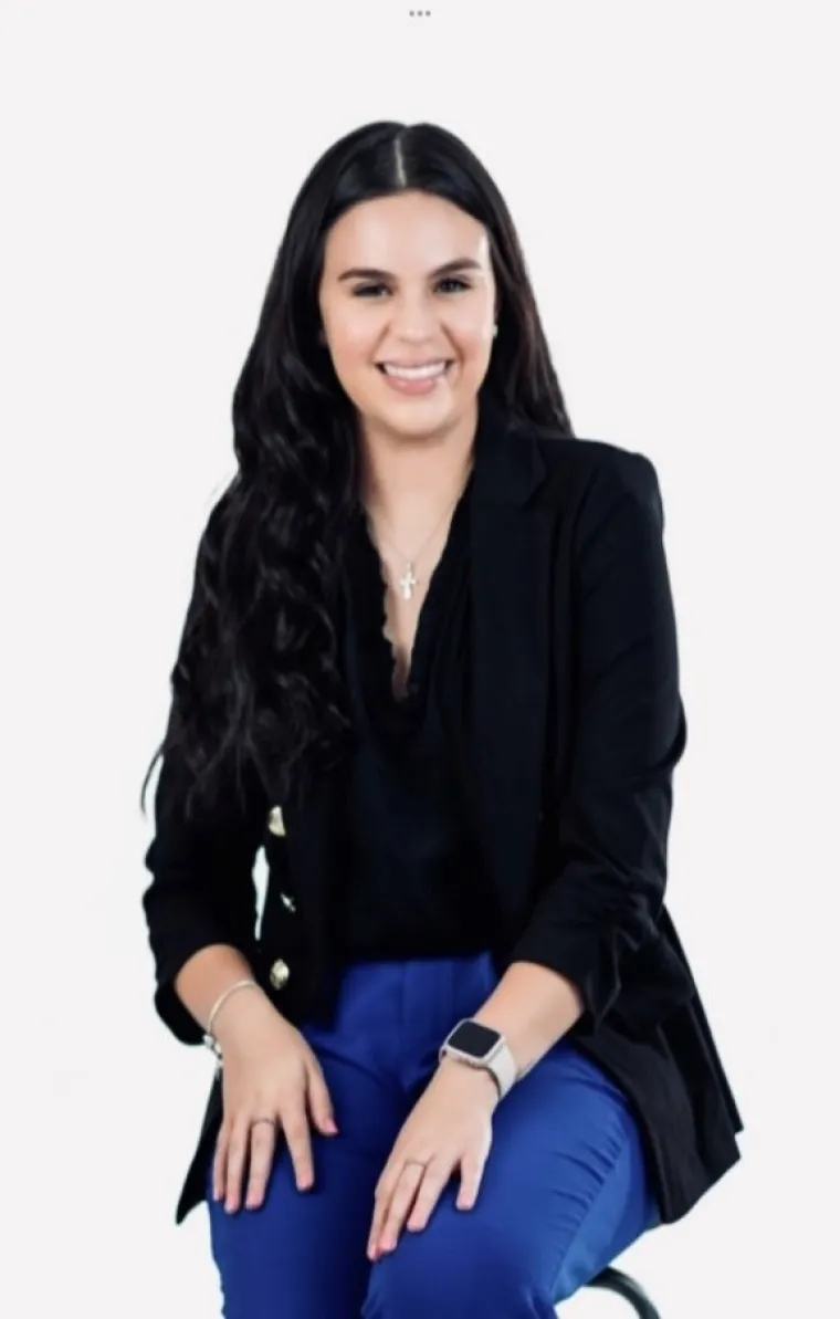 Adriana has long black hair and it smiling at the camera and wearing a black blazer and blue pants.