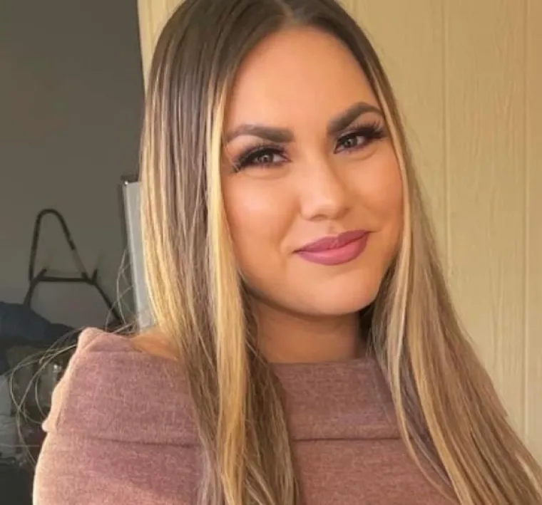 Alexis, a woman of color with long brunette hair with blonde highlights, is standing in front of a white background. She wears a brown sweater and smiles at the camera. 