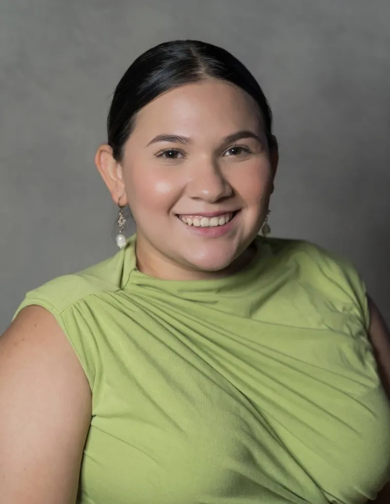 Paulette is sitting in front of a gray backdrop. She has dark hair and is wearing long pearl earrings with a lime green top. Paulette is smiling and looking at the camera.