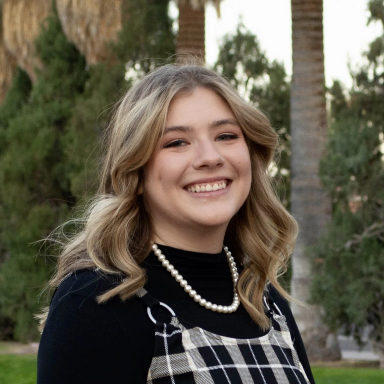 Photo of woman with blonde shoulder length hair, wearing a pearl necklace and a black shirt with black and white patterned overalls, she is smiling at the camera