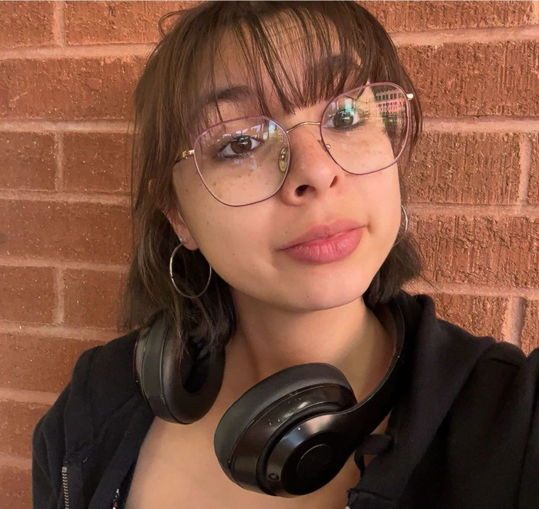 photo of Ariana with brown hair and bangs, wearing a black sweater and glasses with rose gold rims, is staring at the camera