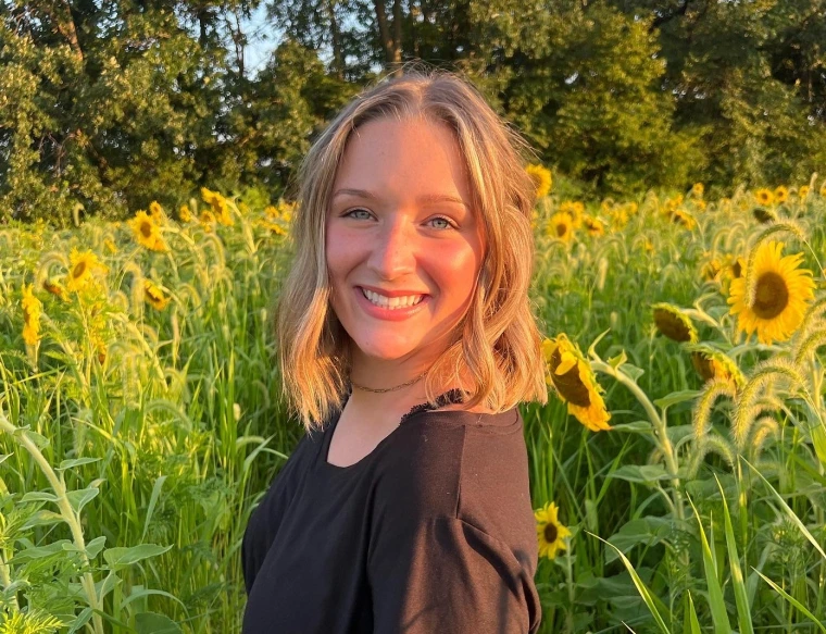woman with short brunette hair, she is wearing a black blouse and standing in front of a sunflower field smiling at the camera