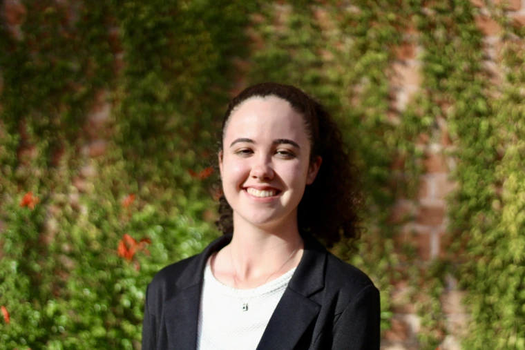 photo of young woman with curly hair in a pony tail, she is wearing a white shirt and black blazer and is smiling at the camera