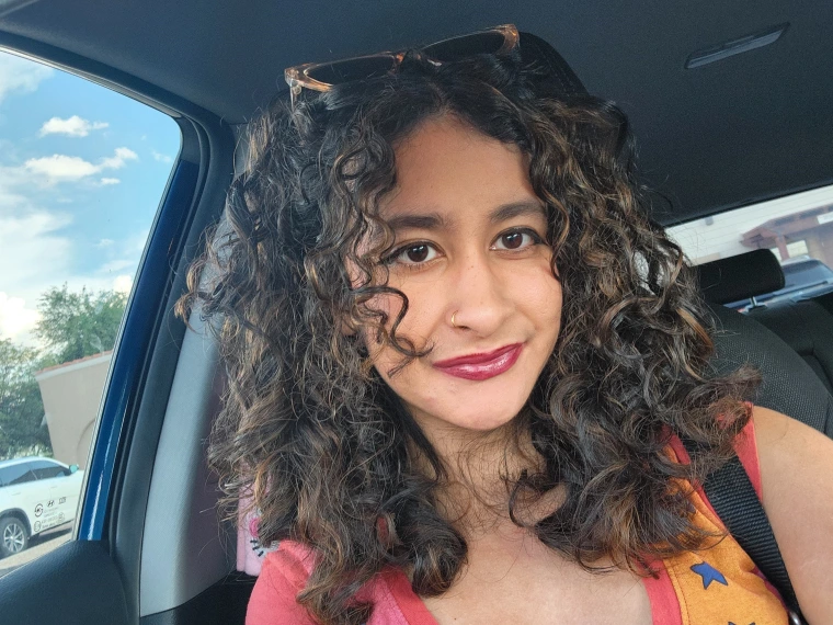 Photo of woman with curly brown hair, wearing a organe and pink shirt and staring at the camera