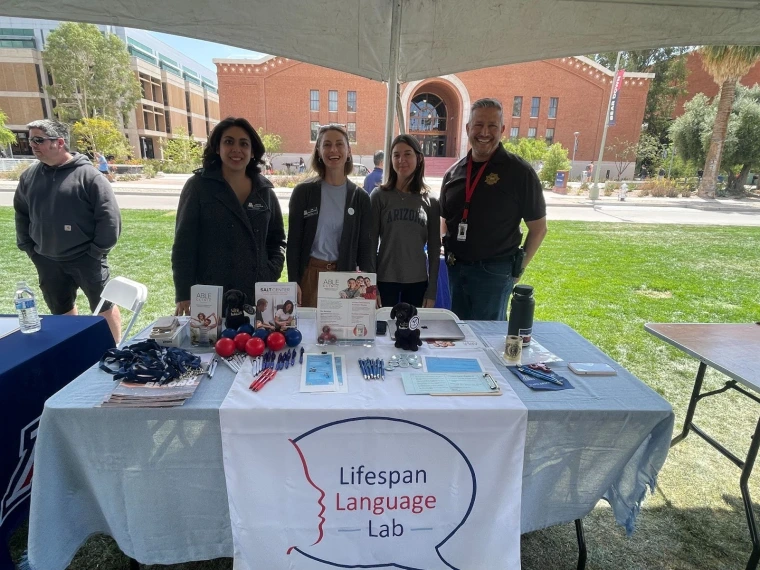  Lifespan Language Lab standing behind their table. The table has fliers, brochures, and assorted swag like pens, stress balls, and lanyards. 