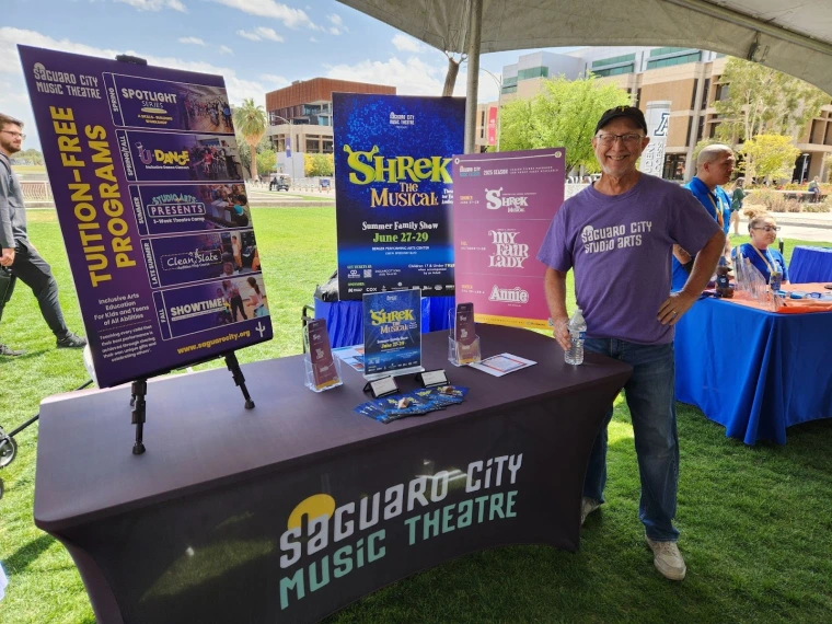 Saguaro City Music Theatre representative standing beside their table. The table has a large poster advertising their production of “Shrek: the Musical” and another poster detailing their other shows in the current season.