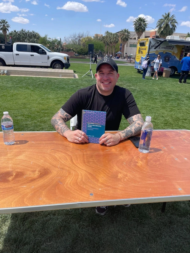 Bradley Irish smiling and sitting at a table, holding a copy of his book “Literary Neurodiversity Studies: Current and Future Directions”.