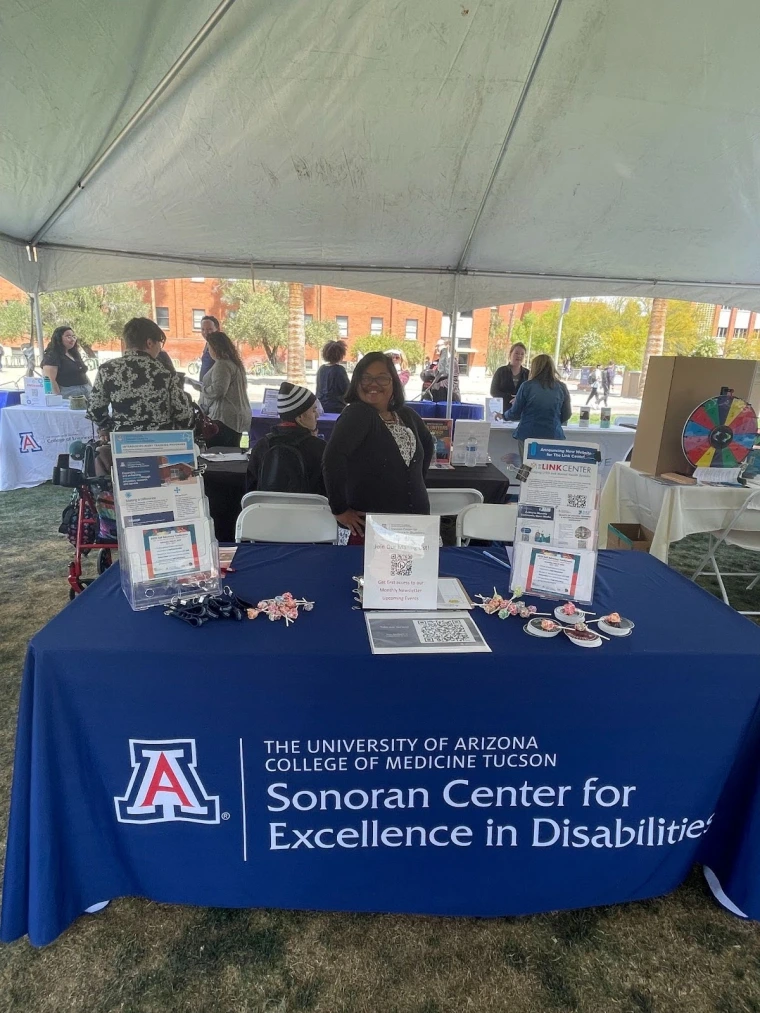Pavleena Madhivanan smiles behind the Sonoran Center table. The table has fliers, a qr code, and assorted candies.