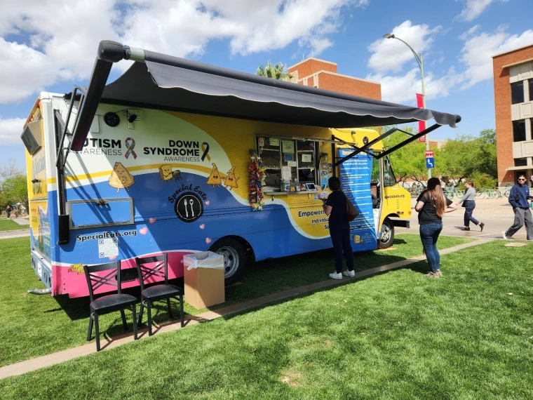 A second Special Eats food truck. The truck is blue and yellow with cartoon illustrations of anthropomorphized nachos, quesadillas and sandwiches, as well as graphics for Down Syndrome and Autism awareness.