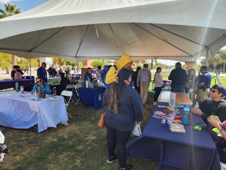  A wide shot of the tabling area at Neurodiversity Day. The tables are on a grassy area under a large tent. Attendees are talking with the various organization representatives at their respective tables.